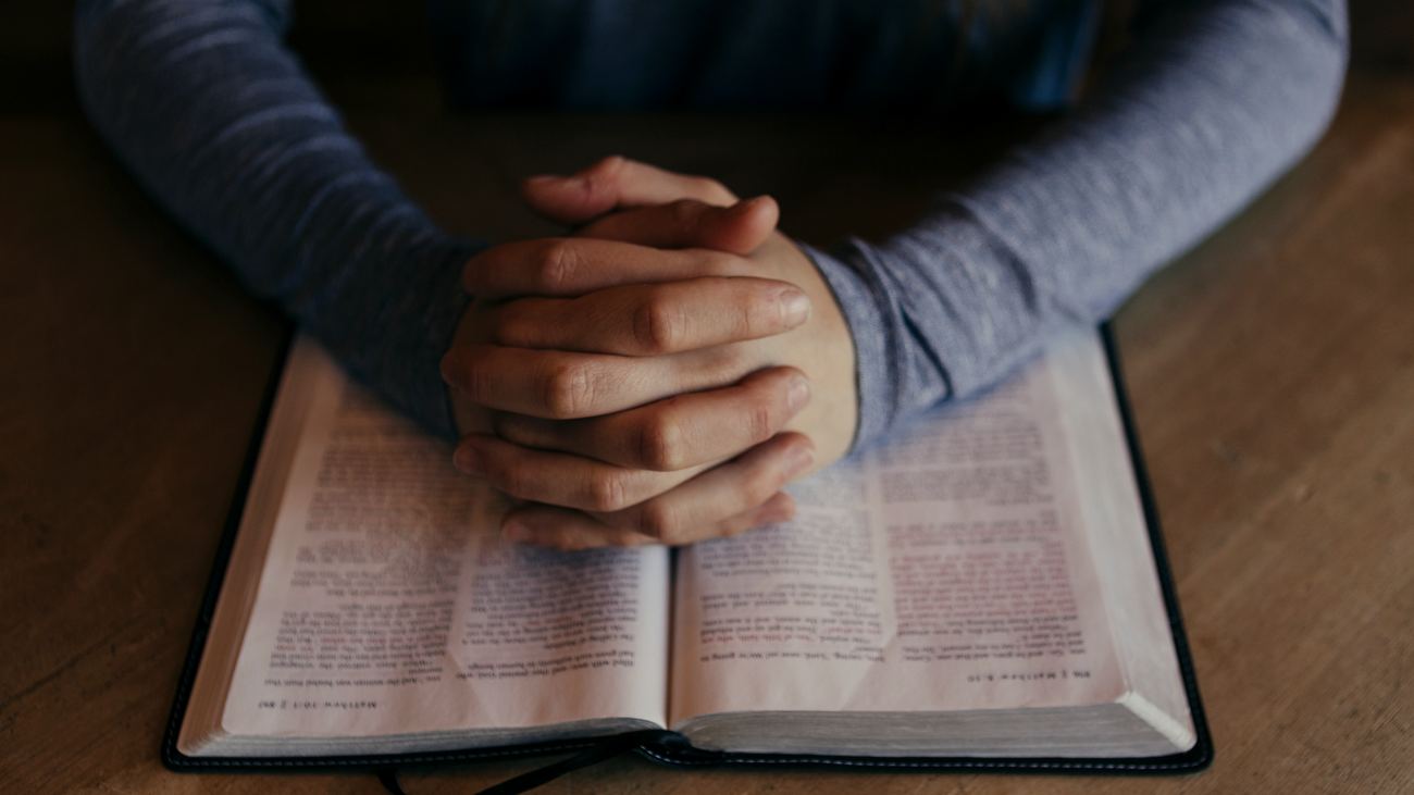 Hands folded on an open Bible resting on a wooden table, in soft window light.