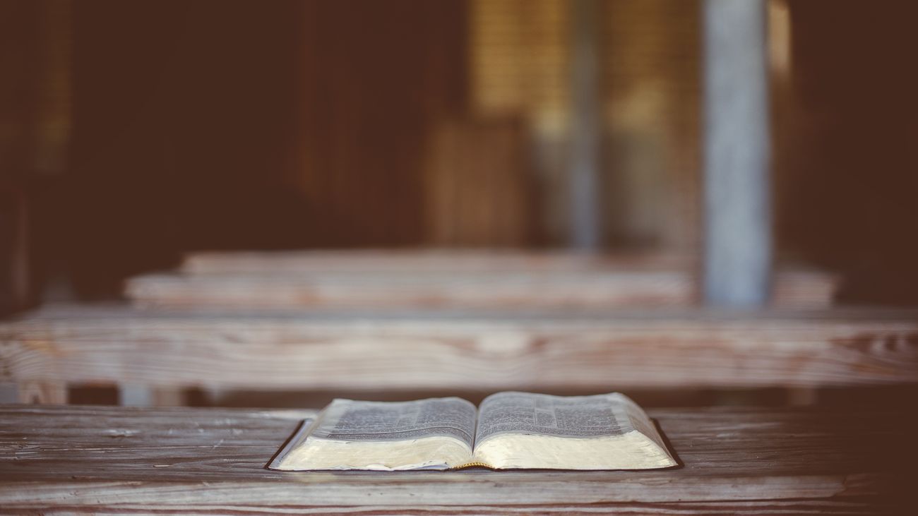 An open Bible lying on a worn wooden pew in a plain, sunlit church.